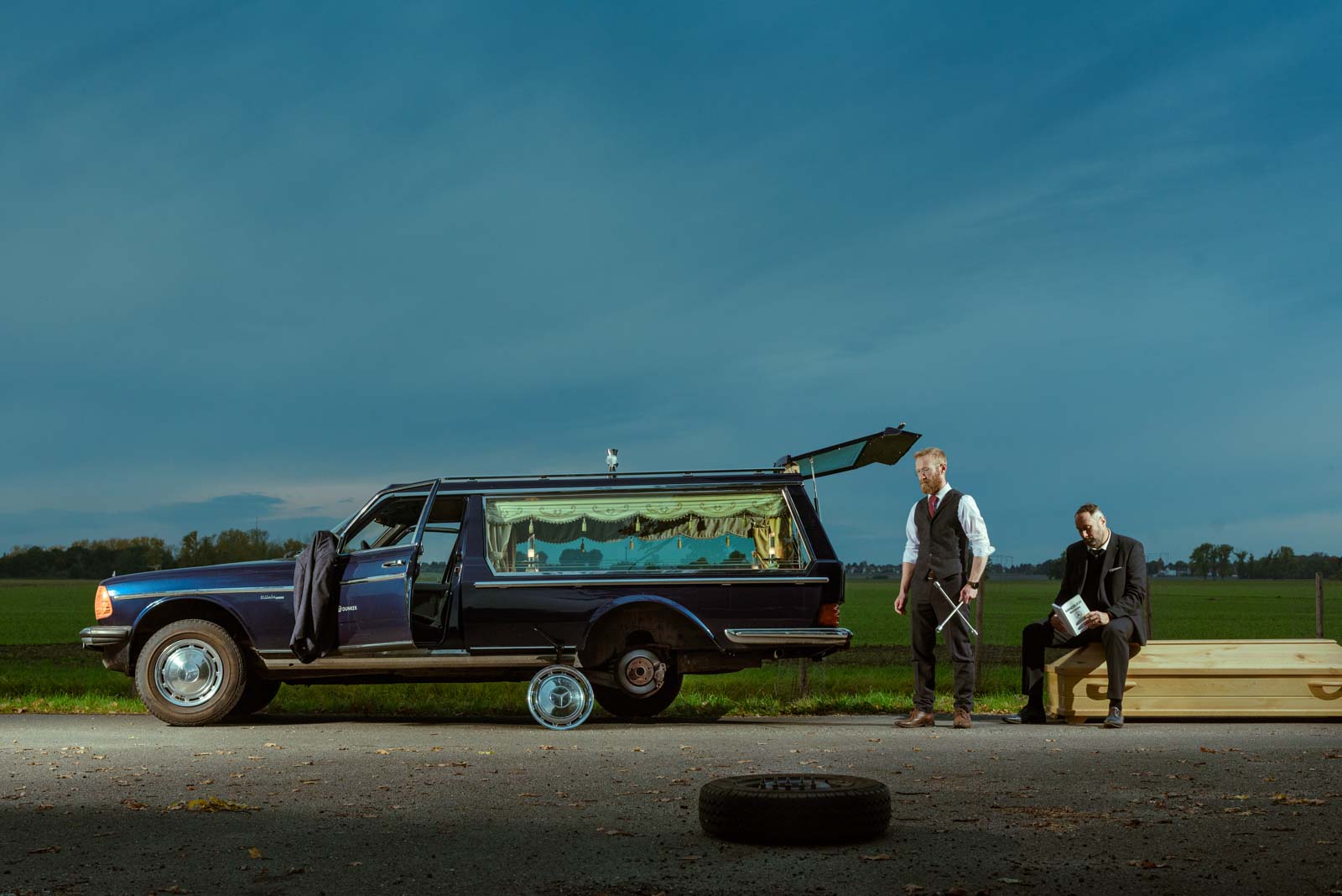 Zwei Männer in Anzügen neben einem alten Bestattungswagen auf einer Landstraße, der hinten geöffnet ist und keinen Hinterreifen hat, während einer sitzt auf einem großen Sarg und der andere steht mit einem Wagenradkreuz in der Hand, im Vordergrund liegt ein Reifen auf dem Boden, im Hintergrund eine weite grüne Wiese unter bewölktem Himmel.