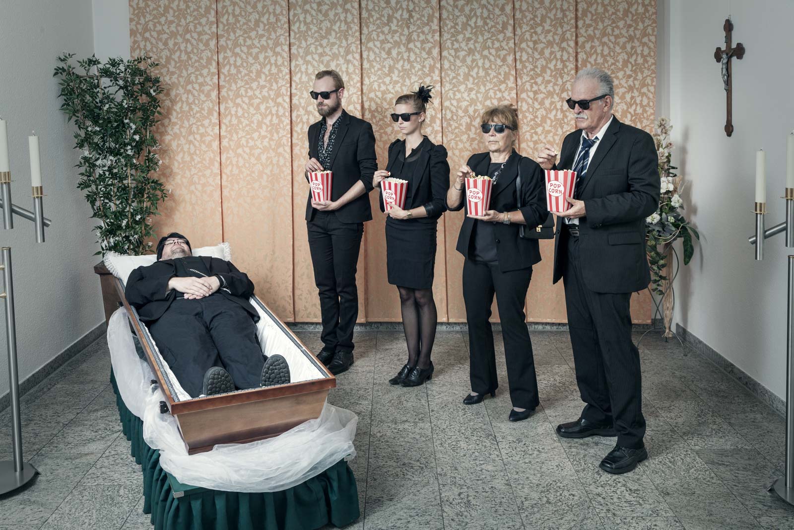 Vier in Schwarz gekleidete Personen mit 3D-Brillen stehen mit Popcorneimern neben einem geöffneten Sarg, in dem ein Mann liegt, die Szene wirkt humorvoll in einem Trauerraum mit Kerzenständern und Kreuz an der Wand.