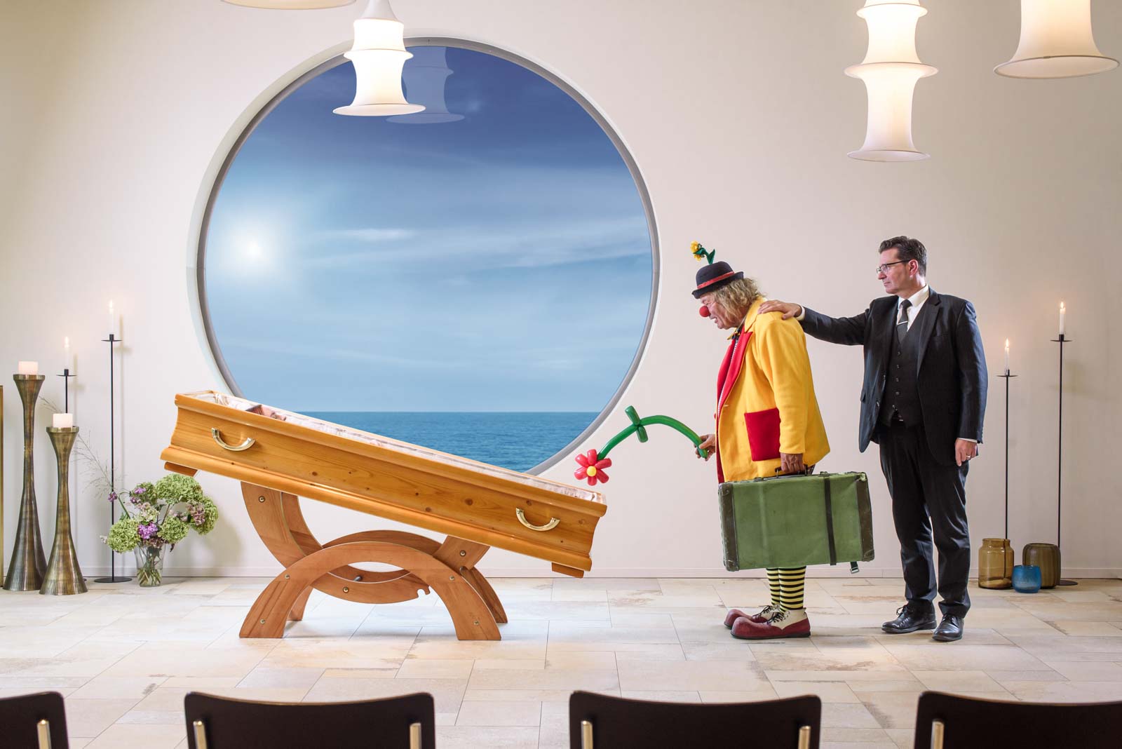 Ein Clown mit roter Nase, gelber Jacke und grünem Koffer steht vor einem Sarg, hält eine Blumenluftballonblume und wird von einem Mann im schwarzen Anzug getröstet, in einem Raum mit runder Fenster mit Blick auf das Meer und mehreren Kerzenständern.