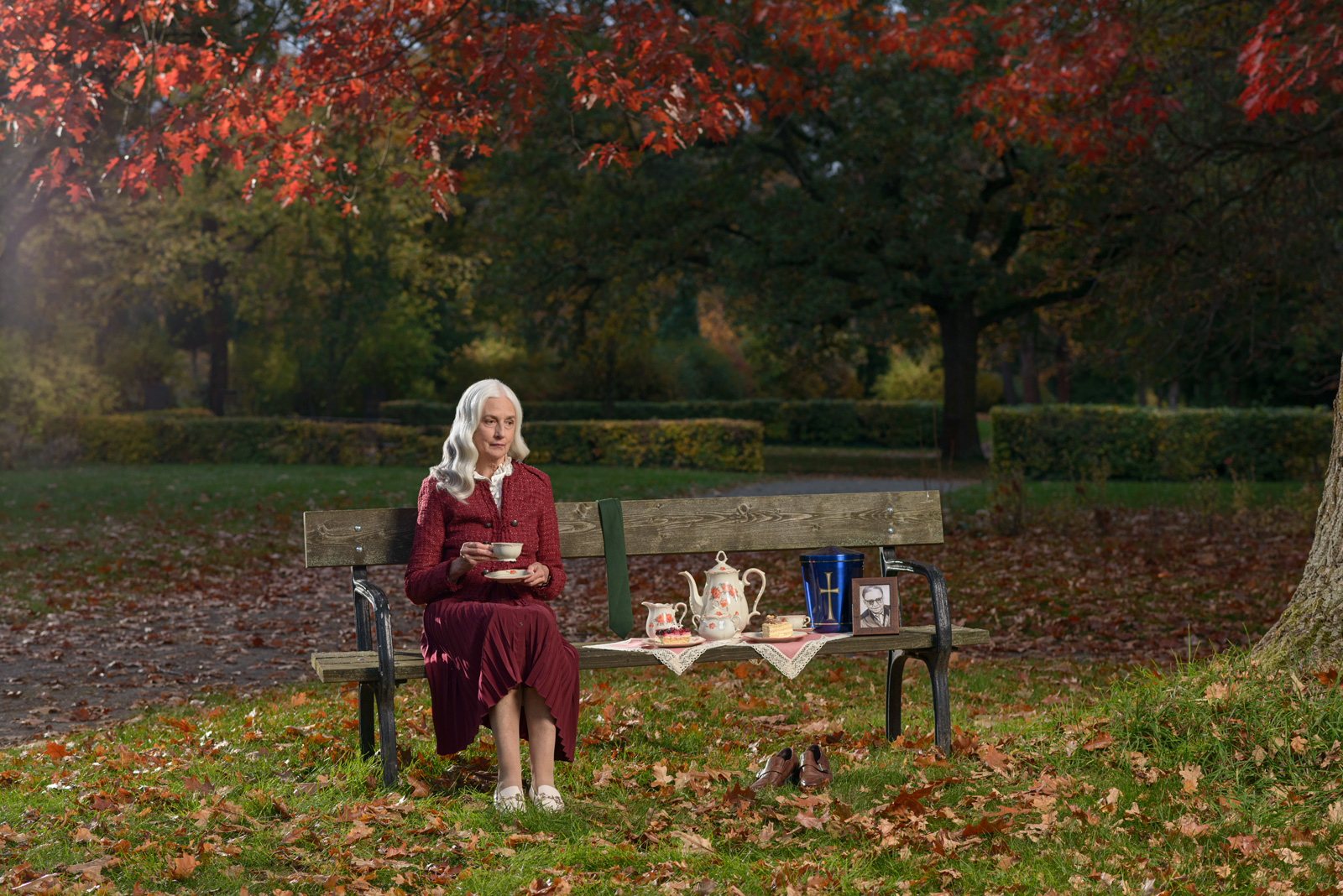 Ältere Frau mit silbergrauen Haaren sitzt auf einer Parkbank im Herbst, umgeben von buntem Laub, trinkt Tee aus einer Tasse, neben ihr auf der Bank eine Teekanne, ein Teller mit Kuchen, eine blaue Urne, ein gerahmtes Foto und ein Paar Schuhe auf dem Gras daneben.
