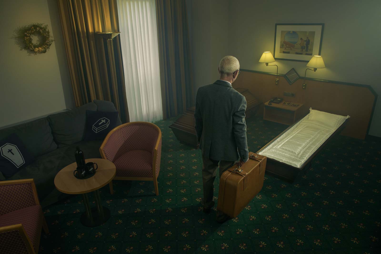 Ein älterer Mann mit einem Koffer steht in einem Hotelzimmer, das mit zwei Särgen, einem Sofa mit Sarg-Motiv-Kissen und einem niedrigen Bett mit weißer Decke ausgestattet ist, beleuchtet von Wandlampen und natürlichem Licht durch ein Fenster mit Vorhängen.